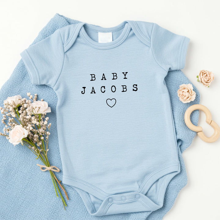 a personalised blue baby vest that says 'Baby Jacobs'. This design features a small heart under the text
