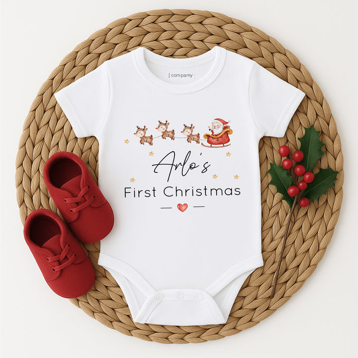 Baby onesie with 'Arla's First Christmas' text, red shoes, and holly leaves on a woven mat.