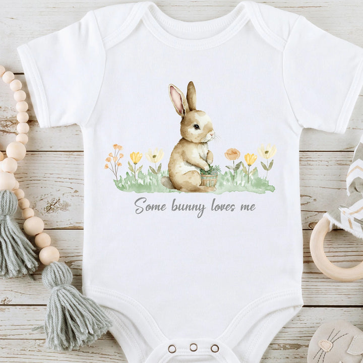 White Easter baby vest that says 'Some bunny loves me'. This design features a brown bunny sitting in flowers.