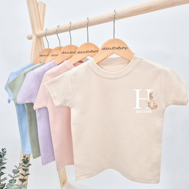 Personalised oatmeal Easter t-shirt that says 'H Harriet'. This design features a rabbbit wearing a beige jacket holding an Easter basket full of eggs