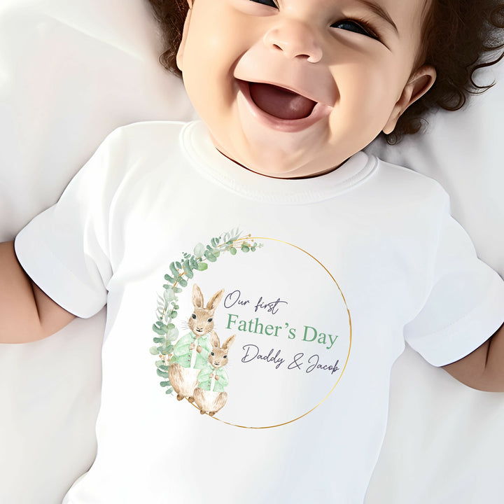 Personalised white Father's Day baby vest that says 'Our First Father's Day Daddy & Jacob'. This design features a gold and green leaf wreath with 2 rabbits wearing green jackets 