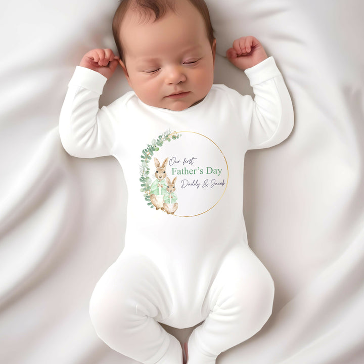 Personalised white Father's Day baby grow/sleepsuit that says 'Our First Father's Day Daddy & Jacob'. This design features a gold and green leaf wreath with 2 rabbits wearing green jackets 