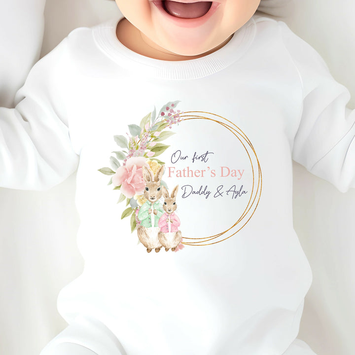 Personalised white Father's Day baby grow/sleepsuit that says 'Our First Father's Day Daddy & Ayla'. This design features a gold leafy wreath with 2 rabbits standing in front. One of the rabbits is wearing a green jacket and the other is wearing a pink jacket