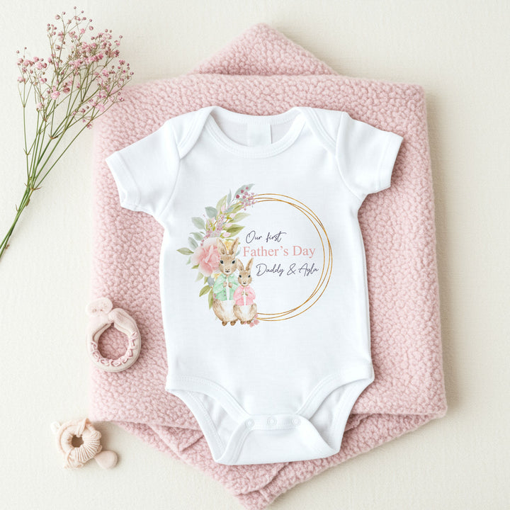 Personalised white Father's Day baby vest that says 'Our First Father's Day Daddy & Ayla'. This design features a gold leafy wreath with 2 rabbits standing in front. One of the rabbits is wearing a green jacket and the other is wearing a pink jacket