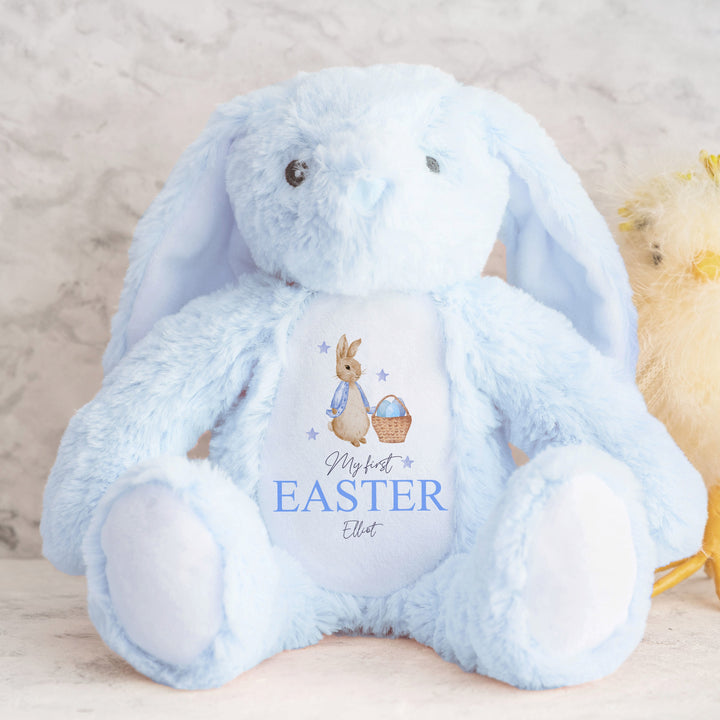 Personalised blue bunny Easter teddy that says 'My First Easter Elliot'. This design features a rabbit wearing a blue jacket holding an Easter basket filled with eggs