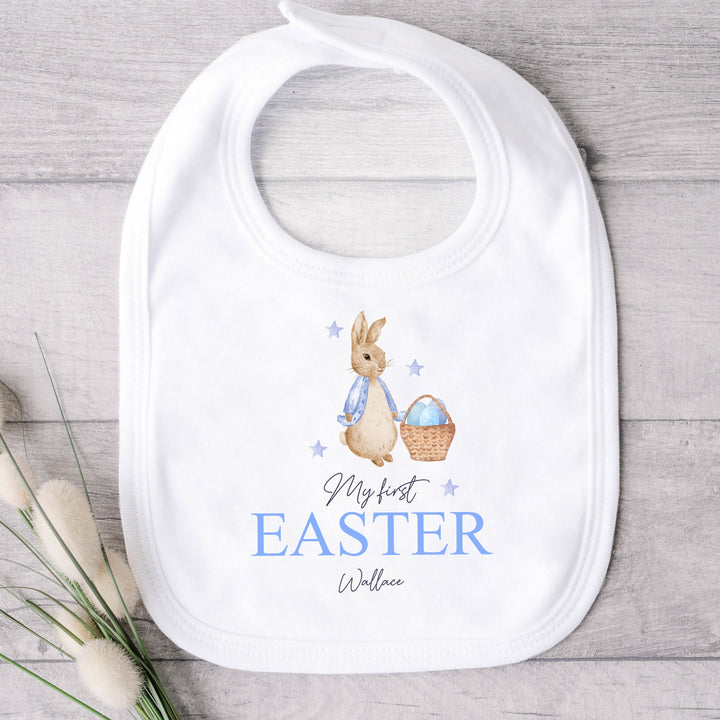 Personalised white Easter baby bib that says 'My First Easter Wallace'. This design features a rabbit with a blue jacket holding an Easter basket with blue stars around it 