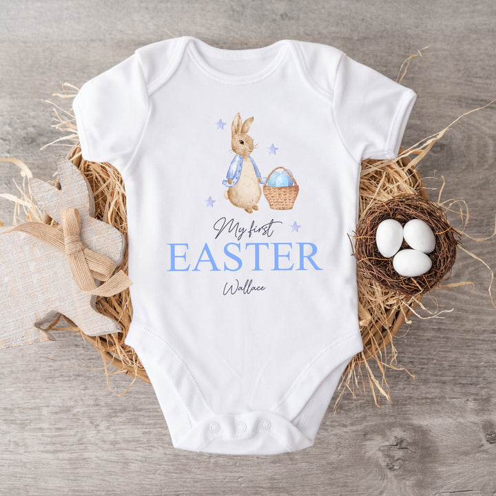 Personalised white Easter baby vest that says 'My First Easter Wallace'. This design features a rabbit with a blue jacket holding an Easter basket with blue stars around it 