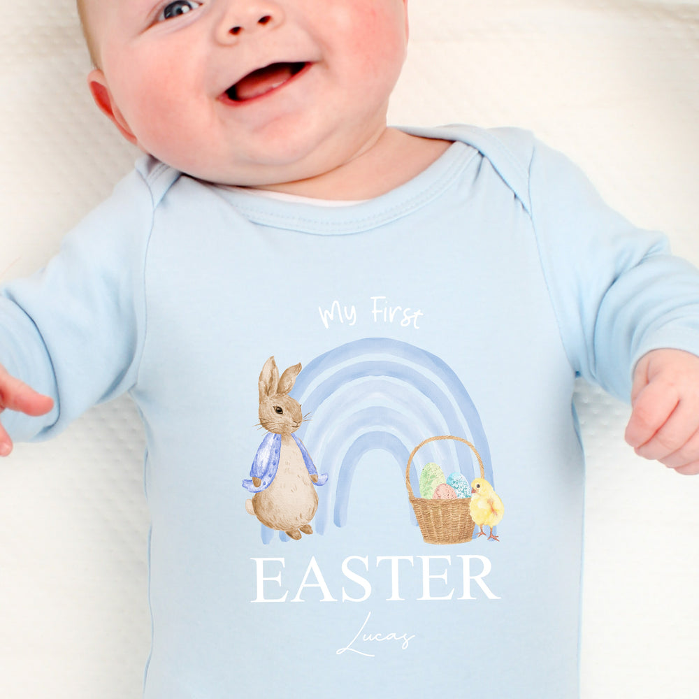 Personalised blue Easter baby grow/sleepsuit that says 'My First Easter Grayson'. This design features a blue rainbow with a rabbit wearing a blue jacket standing one side and an Easter basket and yellow chick the other