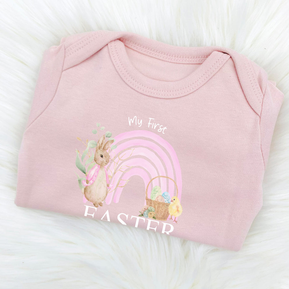 Personalised pink Easter baby grow/sleepsuit that says 'My First Easter Gracie'. This design features a pink rainbow and a rabbit wearing a pink jacket 