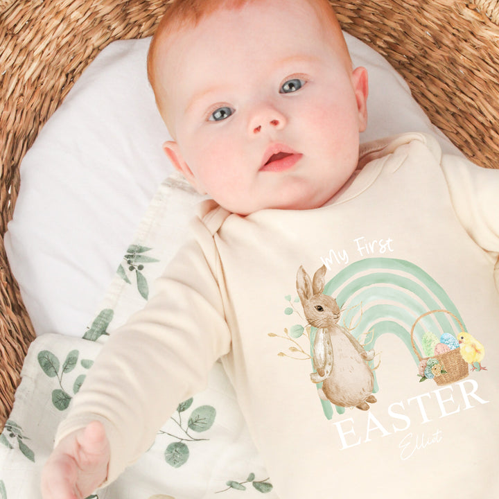 Personalised beige Easter baby grow/sleepsuit that says 'My first Easter Elliot'. This design features a sage green rainbow with a rabbit wearing a beige jacket on one side an Easter basket filled with eggs