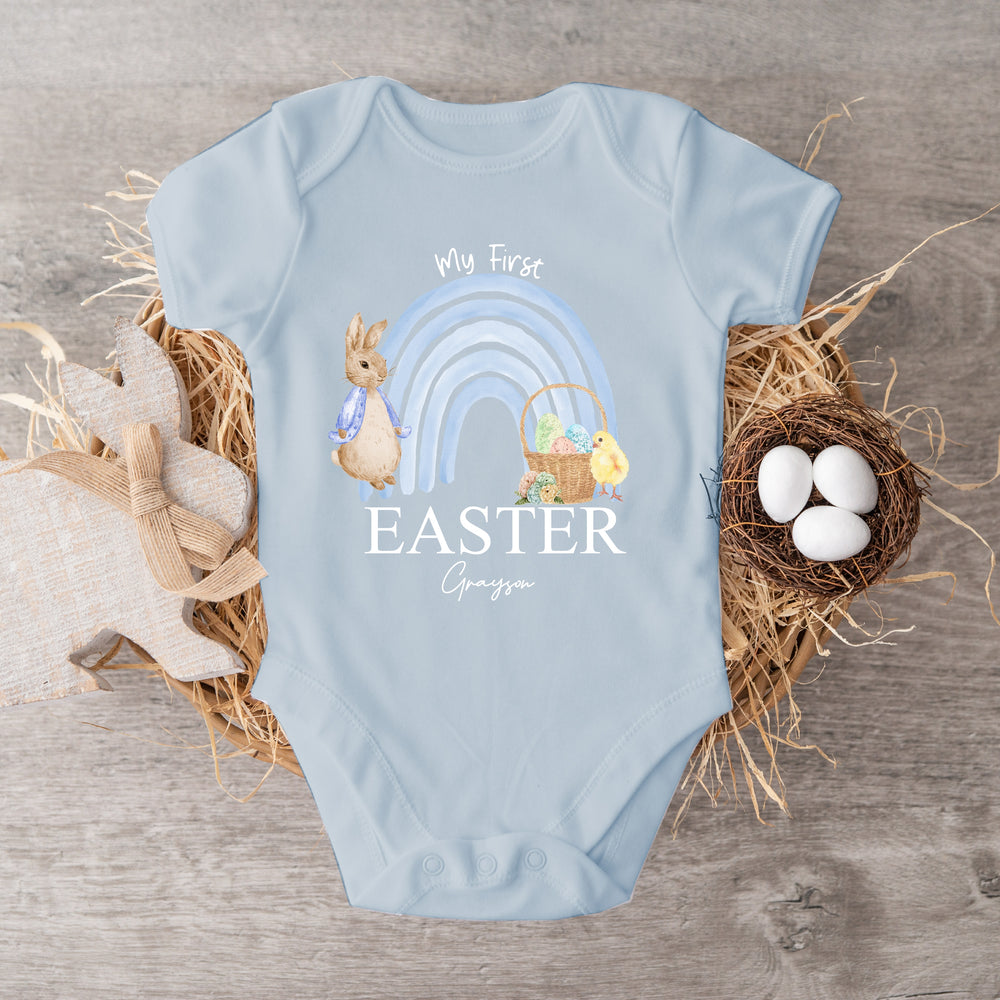 Personalised blue Easter vest that says 'My First Easter Grayson'. This design features a blue rainbow with a rabbit wearing a blue jacket standing one side and an Easter basket and yellow chick the other