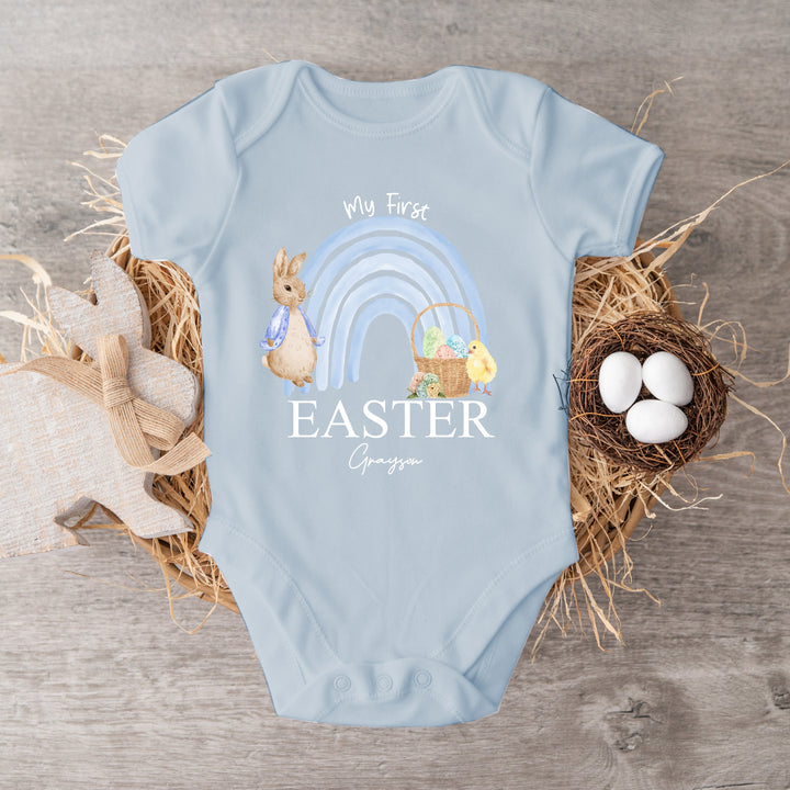 Personalised blue Easter vest that says 'My First Easter Grayson'. This design features a blue rainbow with a rabbit wearing a blue jacket standing one side and an Easter basket and yellow chick the other