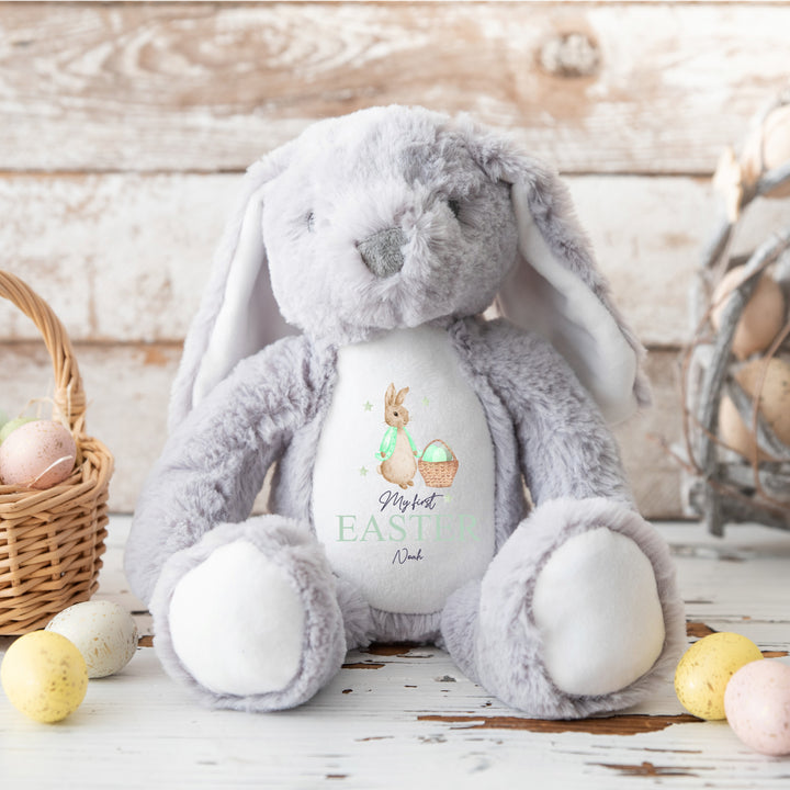 Personalised grey bunny Easter teddy that says 'My First Easter Noah'. This design features a rabbit wearing a green jacket next to a basket full of Easter eggs