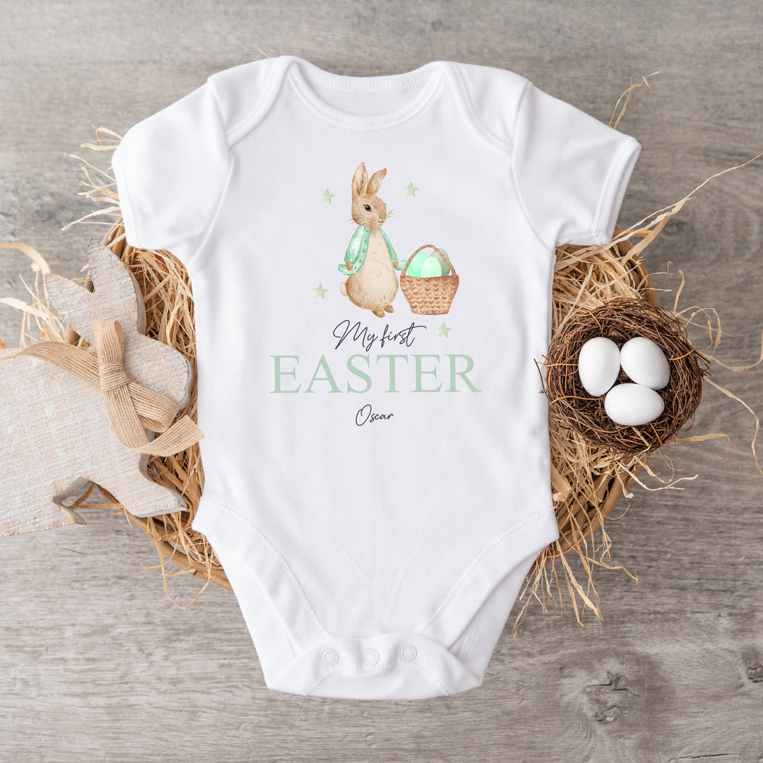 Personalised white Easter baby vest that says 'My First Easter Oscar'. This design features a rabbit wearing a green jacket next to an Easter basket full of eggs