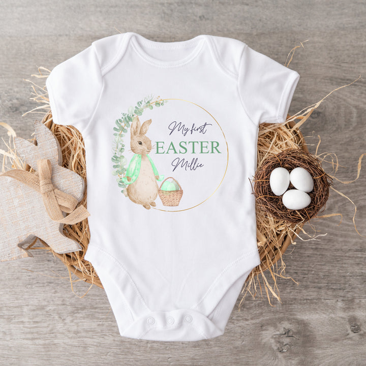 Personalised white Easter baby vest that says 'My First Easter Millie'. This design features a gold and green wreath with a rabbit in a green jacket next to an Easter basket