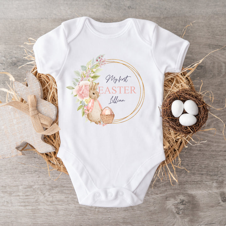 Personalised white Easter baby vest that says 'My First Easter Lillian'. This design features a gold and pink wreath with a rabbit wearing a pink jacket and an Easter basket full with pink eggs