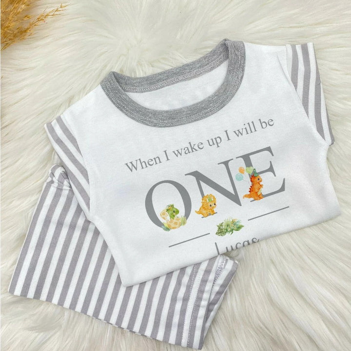 A pair of grey and white stripe children's birthday pyjamas with a dinosaur design saying "When I wake up I will be One" with the name "Lucas".