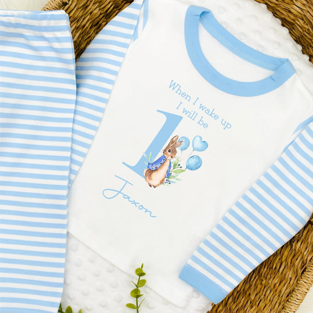 A pair of printed blue striped pyjamas. The design features a rabbit with text saying "when I wake up I will be 1". A name can be added at the bottom of the design.