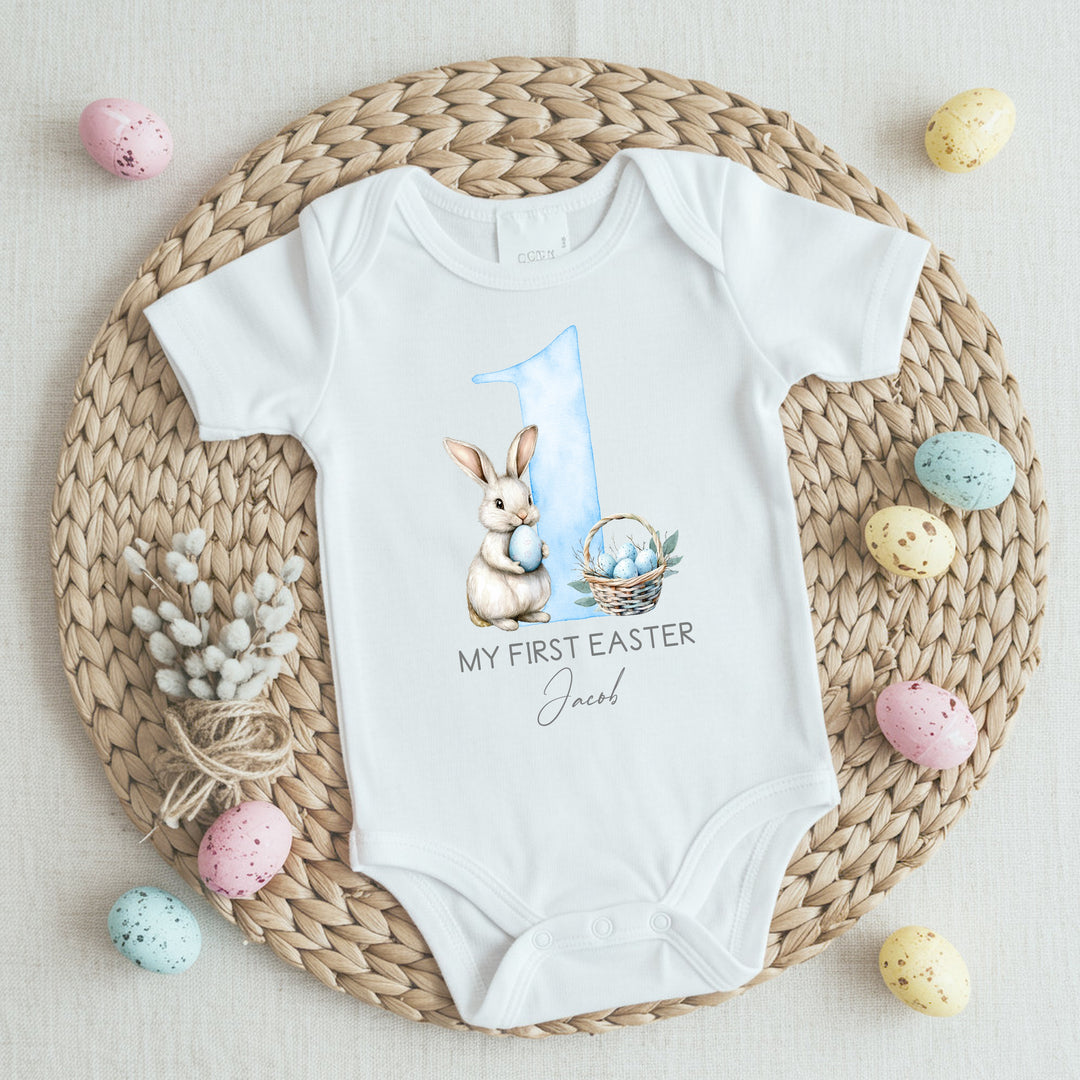 Personalised white Easter baby vest that says 'My First Easter Jacob'. This design features a big blue 1 with a white bunny holding a blue Easter egg