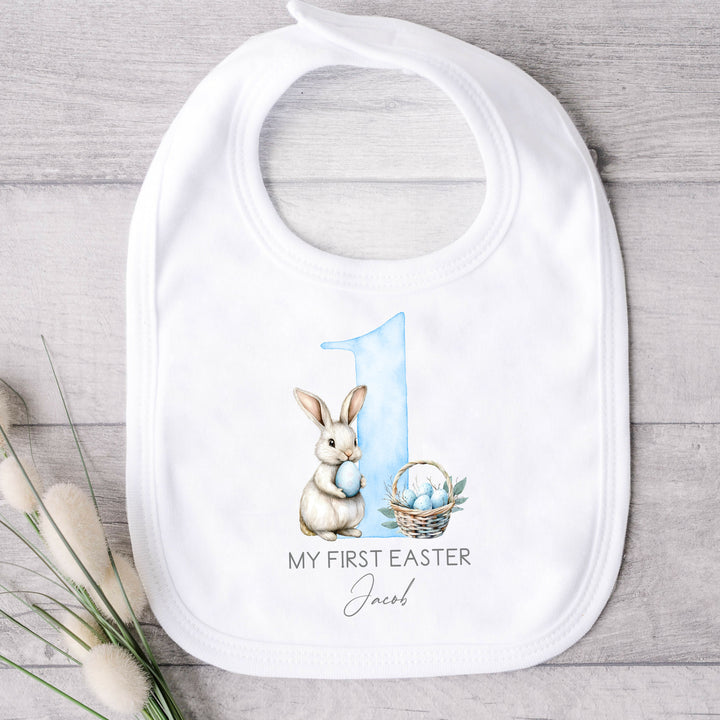 Personalised white Easter baby bib that says 'My First Easter Jacob'. This design features a big blue 1 with a white bunny holding a blue Easter egg