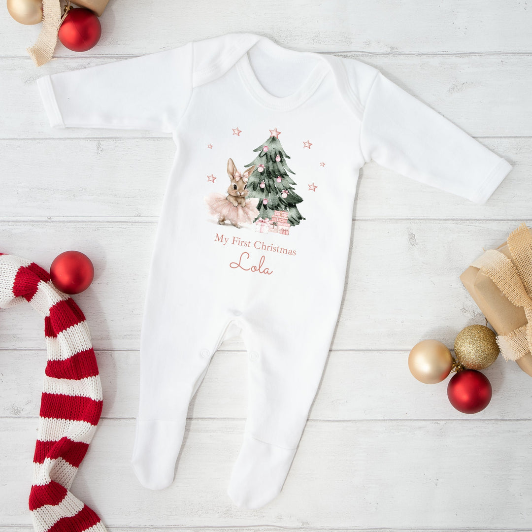 White baby onesie with Christmas design and text, surrounded by festive decorations on a light wooden floor.