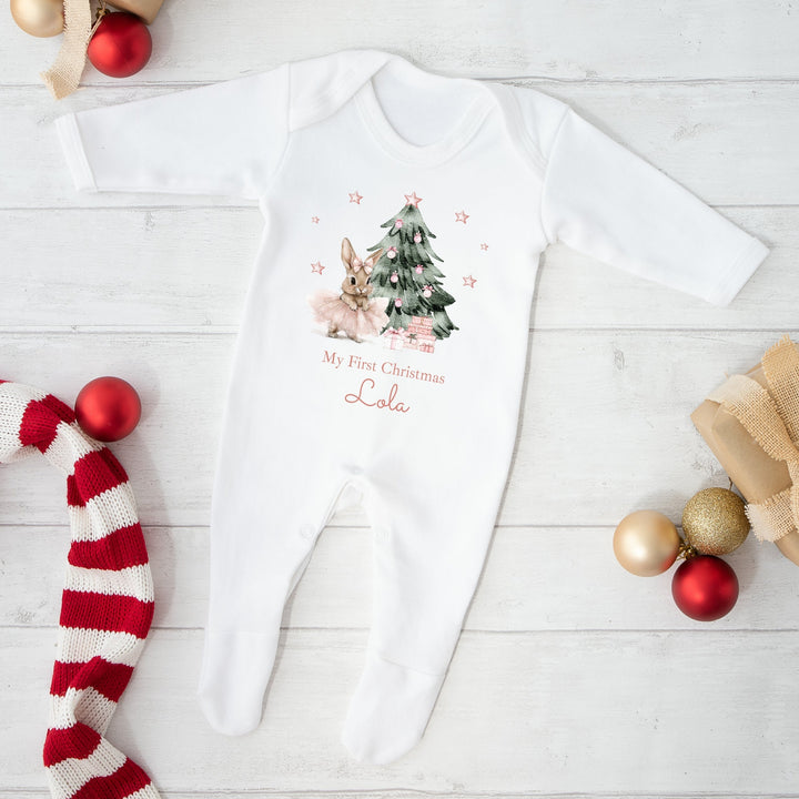 White baby onesie with Christmas design and text, surrounded by festive decorations on a light wooden floor.
